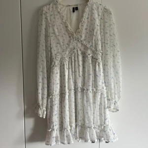 Vit blommig kortklänning Vero Moda - Supersöt vit kortklänning från Vero Moda med små blå blommor, volangdetaljer och v-ringning. Klänningen har långa ärmar med resår vid handlederna och omlottkänsla upptill. Perfekt för sommarens alla tillfällen!