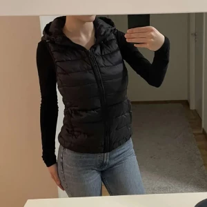 Svart dunväst med huva - Säljer en svart dunväst med huva och dragkedja framtill. Västen är quiltad med breda kanaler och har en sportig look. Perfekt att slänga över en hoodie eller långärmad tröja för extra värme. Snygg och enkel att matcha till jeans eller träningskläder.
