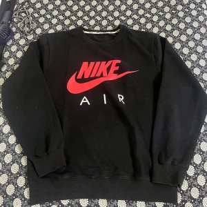 Svart Nike Air sweatshirt med röd logga - Svart sweatshirt från Nike med stor röd Nike Air-logga framtill. Tröjan har rund hals, långa ärmar och ribbade muddar vid ärmslut och nederkant. Perfekt för en sportig och avslappnad stil. Lite smutsig men allt går bort i tvätten.