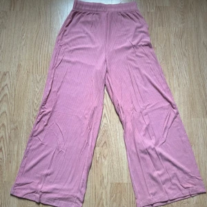 Puderrosa pull-on byxa - Säljer ett par puderrosa ribbade byxor med croppad längd och resår i midjan. Byxorna har en loose fit och är tillverkade i ett mjukt, stretchigt material som känns skönt mot huden. Perfekta för en chill och avslappnad stil