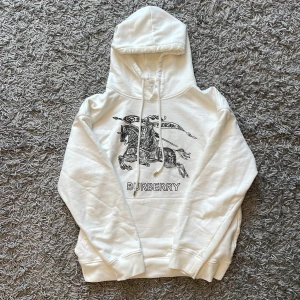 Burberry hoodie - Säljer en vit hoodie från Burberry med ikoniskt svart tryck framtill som visar en riddare på häst och texten BURBERRY. Tröjan har huva med snörning och ribbade muddar. Perfekt för dig som gillar stilrena designer med lyxig känsla.