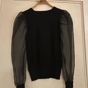 Svart tröja med transparenta puffärmar - Snygg svart tröja med statement-puffärmar i transparent material. Kroppen är i mjukare tyg medan ärmarna är tunna och ger en cool kontrast. Perfekt för dig som vill sticka ut lite extra med din outfit.