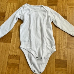 Vit långärmad body från Dofi Baby - En basic vit långärmad body från Dofi Baby i mjuk bomull. Bodyn har praktiska tryckknappar i grenen för enkel på- och avklädning. Perfekt att ha som lager under andra plagg under kyligare dagar.