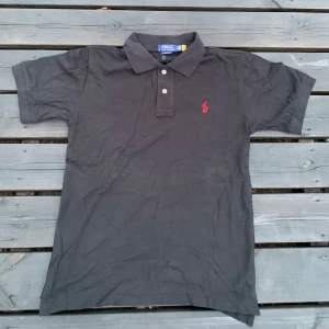 Svart pikétröja Polo Ralph Lauren S - Svart pikétröja från Polo Ralph Lauren i slim fit med klassisk krage och två knappar framtill. Tröjan har röd broderad logga på bröstet och är tillverkad i mjuk bomull. Perfekt för en clean och stilren look.
