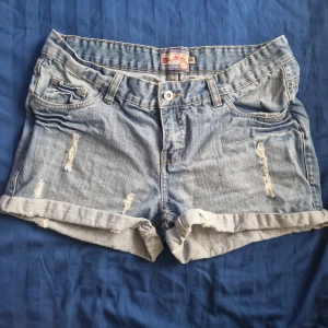 blå jeansshorts från Miss Milla - Snygga blå jeansshorts från Miss Milla med slitna detaljer och uppvikta benslut. Bakfickan har coola pärl detaljer i vitt och blått. Klassisk femficksmodell och normal passform, perfekta för sommardagar. Obs, lite mörkare irl än på bilderna
