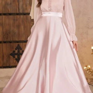 Ljusrosa långklänning med pärldetaljer - Elegant ljusrosa långklänning i glansigt satinliknande material med transparenta långärmar och v-ringning dekorerad med pärlor. Klänningen har markerad midja med bred satinbård och ett svepande, utsvängt kjolparti. Perfekt för bal eller fest.