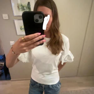 Vit blus med volangärmar - Supersöt vit blus med korta volangärmar och broderade detaljer. Toppens ärmar har en romantisk känsla med hålbroderi och lätt rynk. Rak passform och rund halsringning, perfekt till jeans eller kjol.