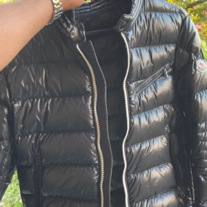 Svart Moncler pufferjacka med dragkedjor - Snygg svart pufferjacka från Moncler med glansig finish och klassisk logga på ärmen. Jackan har flera dragkedjeförsedda fickor och en rak passform. Perfekt för dig som vill ha en stilren och varm jacka med exklusiv känsla.