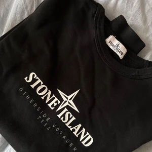 Svart Stone Island sweatshirt - Svart sweatshirt från Stone Island med vit logga och text framtill. Klassisk rund halsringning och långa ärmar med ribbade muddar. Tröjan är i den största junior storleken men sitter som en S/M