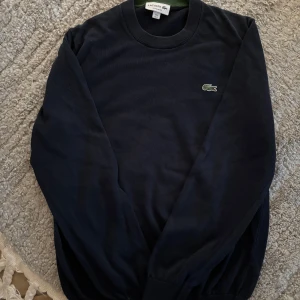 Mörkblå tröja från Lacoste - Säljer en klassisk mörkblå tröja från Lacoste med rund halsringning och det ikoniska broderade krokodilloggan på bröstet. Tröjan har långa ärmar och ribbade muddar vid ärmslut och nederkant. Perfekt för dig som gillar stilren och tidlös design.