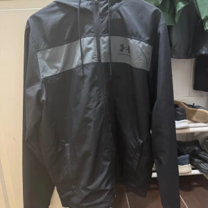 Svart vindjacka från Under Armour L - Svart vindjacka från Under Armour med grått panelparti över bröstet och logga. Jackan har huva med snörning, dragkedja framtill och två sidofickor. Lätt och sportig känsla, perfekt för blåsiga dagar. Passform loose för en avslappnad stil. Möts gärna mvh 