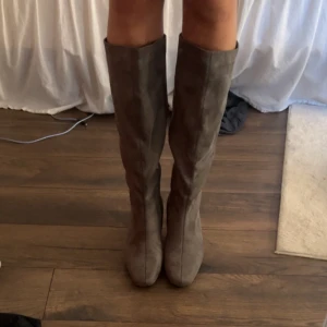 Grå mockastövlar med låg klack - Snygga knähöga stövlar i grå mocka med låg klack och rund tå. Perfekta för att lyfta din outfit med en stilren och trendig touch. Stövlarna har en klassisk siluett och är lätta att matcha till olika looks.