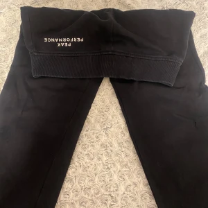 Svarta mjukisbyxor Peak Performance - Svarta mjukisbyxor från Peak Performance med resår och snörning i midjan. Byxorna har ribbade muddar vid bensluten och är tillverkade i mjukt material som passar perfekt för chill eller träning. Diskret logga på ena benet.