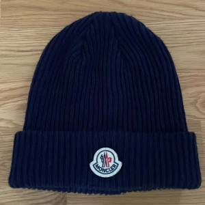 Moncler mössa - Snygg mörkblå ribbstickad mössa från Moncler med klassisk uppvikt kant och broderad logotyp framtill. Perfekt accessoar för kalla dagar och ger en clean streetstyle-vibe.