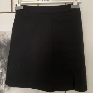 Svart kortkjol från BikBok, stl 34 - Enkel och stilren svart kortkjol från BikBok i storlek 34. Kjolen har en liten slits framtill och dragkedja bak. Perfekt för dig som gillar minimalistisk stil och vill ha en klassisk kjol i garderoben.