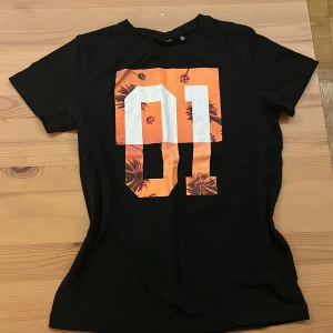 Svart t-shirt med orange print REACTION - Svart tränings t-shirt från REACTION med stort orange och vitt tryck av siffran 01 på bröstet, där det orangea har palmmotiv. Klassisk rund hals och korta ärmar. Perfekt för en cool och avslappnad stil vid träning.