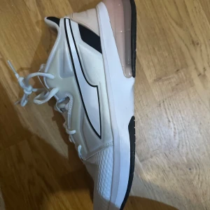 Vita sneakers från Puma strl 41 - Snygga vita Puma sneakers med svarta detaljer och rosa transparent sula. Skorna har snörning, meshpartier för andning och robust platt sula med PUMAGRIP för extra grepp. Perfekta för dig som gillar sportig och modern stil.