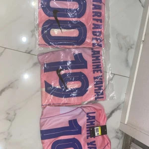 Rosa Nike fotbollströja Lamine Yamal - Snygg rosa fotbollströja från Nike med Lamine Yamal och nummer 19 tryckt i blått på ryggen. Tröjan har korta ärmar och sportig passform, perfekt för dig som vill sticka ut på planen eller visa support för din favoritspelare.