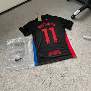 FC Barcelona x Nike Raphinha tröja - Säljer en svart FC Barcelona fotbollströja från Nike med Raphinha och nummer 11 på ryggen. Tröjan har röda och blå sidopaneler, klubbmärke på bröstet och Travis Scott-inspirerad grafitti-text på framsidan. Tillverkad i lätt och ventilerande material.