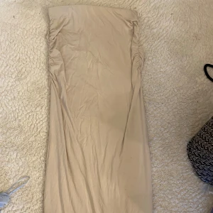 Beige axelbandslös långklänning - En stilren beige långklänning med axelbandslös design och rynkade sidor upptill. Klänningen har en rak och enkel siluett, perfekt för dig som gillar minimalistisk stil. Materialet känns mjukt och följsamt mot kroppen.