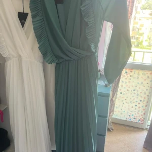 Grön långklänning med volangdetaljer - Snygg grön långklänning med plisserad kjol och omlottliv. Klänningen har långa ärmar i ett lätt, flowy material och volangdetaljer längs urringningen. Perfekt för dig som vill sticka ut med en elegant och trendig look. Det finns en vita klänning på samma modell,och priset gäller på ett klänning, och vi kan diskutera priset. 