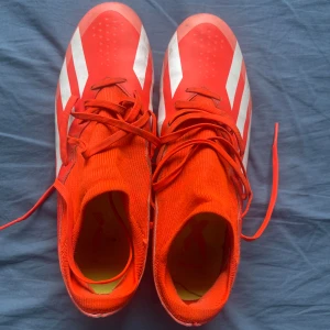 Adidas X röda fotbollsskor med snörning - Snygga röda Adidas X fotbollsskor med vita detaljer och klassiska tre ränder på sidorna. Skorna har snörning, plös i stickad textil och dobbar i gult och vitt för bra grepp på planen. Perfekta för dig som vill sticka ut på matchen.