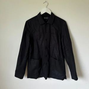 Svart overshirt från Riley i linneblandning - Stilren svart overshirt från Riley i linneblandning. Jackan har klassisk krage, knäppning framtill och stora fickor. Perfekt för lager-på-lager och funkar till många olika outfits. Enkel och clean look med lång ärm och avslappnad passform. Artnr: A346