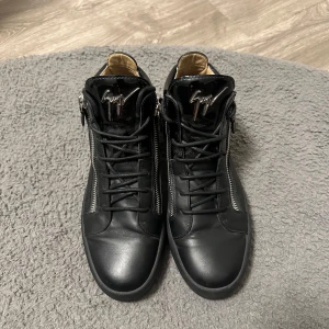 Svarta sneakers från Giuseppe Zanotti - Snygga svarta sneakers från Giuseppe Zanotti i skinn med glansiga detaljer och silverfärgade dragkedjor på sidorna. Skorna har rund tå, snörning och en tjock platt sula. Ikonisk logga på plösen och cool italiensk design.