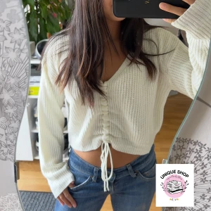 Vit croppad stickad tröja med snörning - Superfin vit stickad tröja med v-ringning och croppad passform. Tröjan har långa ärmar och snygg snörning framtill som ger en cool detalj. Perfekt att styla med jeans för en chill look.