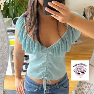 Ljusblå volangtopp med snörning - Supersöt ljusblå topp med volang runt v-ringningen och snörning framtill. Toppen är croppad och har korta ärmar med plisserad struktur. Perfekt för dig som gillar romantisk och trendig stil.