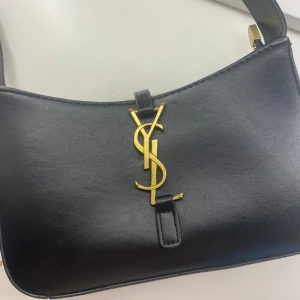 Svart väska från Yves Saint Laurent - Den finaste och populäraste svarta axelväskan från YSL i slätt skinn med ikoniskt guldigt YSL-emblem framtill. Väskan har en stilren, avlång form och justerbart axelband med gulddetaljer. Stängs med dragkedja upptill och passar perfekt till alla evenemang!! 