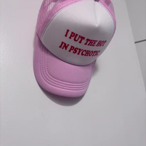 Rosa och vit truckerkeps med text - Snygg truckerkeps i rosa och vitt med röd texten 'I PUT THE HOT IN PSYCHOTIC' framtill. Kepsen har mesh-bakstycke för extra ventilation och justerbar knäppning bak. Perfekt statement-piece för dig som vill sticka ut.