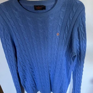 Blå kabelstickad tröja från Morris - Kabelstickad blå tröja från Morris i 100% pima bomull. Klassisk rund halsringning, ribbade muddar och diskret broderad logga på bröstet. Perfekt för dig som gillar stilrena och tidlösa plagg med en preppy vibe.
