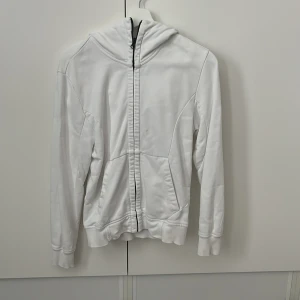 Vit hoodiejacka med goggles C.P. Company - Säljer en vit hoodiejacka från C.P. Company med ikoniska goggles i huvan. Jackan har dragkedja framtill, två fickor och ribbade muddar. Tillverkad i mjukt bomullsmaterial och har en clean, sportig look som sticker ut.