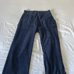Mörkblå mjukisbyxor Juicy Couture XS - Mörkblå mjukisbyxor från Juicy Couture med snörning i midjan, två stora fickor fram och bakfickor med knappar. Byxorna är i mjukt material och har en avslappnad passform, perfekta för chill dagar.