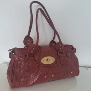 Röd handväska i skinn med guldiga detaljer - Säljer en snygg röd handväska i skinn med krokopräglad yta och guldiga metalldetaljer. Mulberry liknande väska