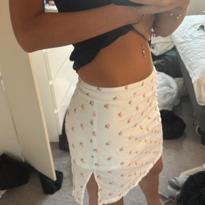 Vit blommig kortkjol med slits - Supersöt vit kortkjol med små rosa blommor och grönbladigt mönster. Kjolen har en liten slits på sidan vilket ger en extra gullig touch. Perfekt för varma dagar och enkel att matcha med en crop top eller linne. 🥰