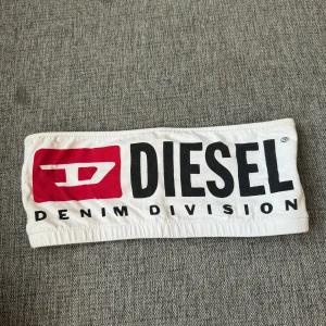 tubtopp från Diesel - Vit bandeau tubtopp från Diesel med stor logga och Denim Division-tryck i svart och rött framtill. Topp i stretchig bomull med elastisk kant för att sitta på plats. Perfekt för dig som vill sticka ut med en ikonisk streetstyle-look.