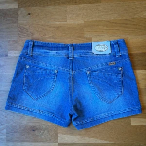 Blå jeansshorts från DND Jeans - Snygga blå jeansshorts från DND Jeans med klassisk femficksdesign, dubbla knappar framtill och uppvikta benslut. Shortsen har bälteshällor och detaljerade sömmar för en cool look. Perfekta för varma dagar och enkel att matcha med allt. Midjemåttet rakt över (liggandes) är ca 38 cm ☺️