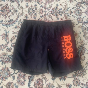 Svarta badshorts från Hugo Boss - Svarta badshorts från Hugo Boss med stor orange logga på ena benet. Shortsen har elastisk midja med snörning och innerbyxa i mesh. Perfekta för stranden eller poolen, med cool kontrast mellan svart tyg och orange detaljer.