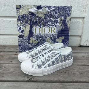 Dior skor  - Snygga Dior sneakers med klassiskt grått och vitt monogrammönster, vita snören och robust vit platt sula med svart stjärndetalj undertill. Ovandel i textil och detaljer i skinn. Perfekta för dig som vill sticka ut med lyxig streetstyle. Inne sulorna e lite skadade och fläck som syns på insidan men inget märks vi användning  