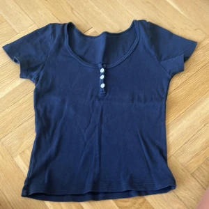 Mörkblå ribbad topp med knappar - Mörkblå ribbad topp . Framtill finns fyra små vita knappar som detalj. Modellen är figurnära och passar perfekt till jeans eller kjol för en enkel och snygg look. den är lite skrynklig, men det är bara att stryka den