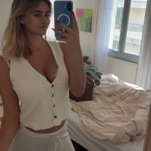 Vit stickad väst med knappar med matchande byxor  - Säljer en vit stickad väst med v-ringning och snygga knappar framtill. Västen är croppad och har en mjuk, ribbad struktur som ger en chill vibe. Perfekt att styla med matchande byxor eller jeans för en clean look.