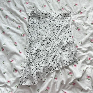 Vit prickig omlottkjol från Kappahl - Supersöt vit omlottkjol från Kappahl med svarta prickar och volangkant. Kjolen har knytband i midjan.