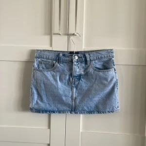 Ljusblå jeanskjol från 157 - Snygg ljusblå jeanskjol från 157 med klassisk femficksdesign, bälteshällor och knappstängning fram. Perfekt för dig som gillar en enkel och tidlös look. Kjolen är kort och har raka linjer för en clean vibe.