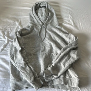 Grå hoodie från H&M med huva - En klassisk ljusgrå hoodie från H&M i storlek S. Tröjan har huva med snörning, magficka och ribbade muddar vid ärmslut och nederkant. Perfekt för en chill och avslappnad stil.