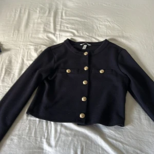 Svart cardigan jacka från H&M med guldknappar - Stilren svart cardigan jacka från H&M med snygga guldknappar framtill och dekorativa fickor. Jackan är croppad och har rund halsringning samt långa ärmar. Perfekt för dig som gillar en klassisk men trendig look.