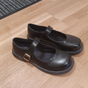 Svarta Mary Jane ballerinaskor - Stilrena svarta ballerinaskor i klassisk Mary Jane-modell med rund tå och justerbart spänne i guld. Skorna har platt sula och är tillverkade i skinnimitation, perfekta för en trendig och tidlös look.