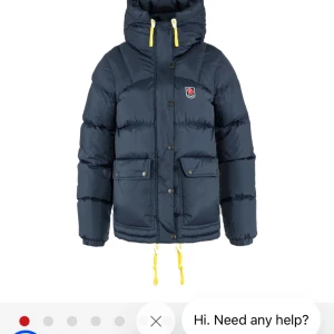 Mörkblå dunjacka från Fjällräven - Snygg mörkblå dunjacka från Fjällräven med gula detaljer på snörningen och huva. Jackan har stora fickor framtill, dragkedja och knappar, samt klassisk Fjällräven-logga på bröstet. Perfekt för kalla dagar och riktigt varm.