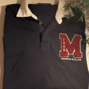 Morris R. Club marinblå pikétröja - Marinblå långärmad pikétröja från Morris R. Club med vit krage och vita ränder på ena ärmen. Stor röd M-patch med broderad text på bröstet. Klassisk rugbyinspirerad stil i bomull, perfekt för dig som gillar preppy vibes.